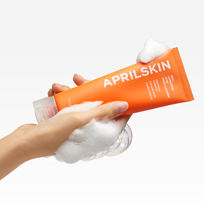 APRILSKIN Real Carrotene Acne Foam Cleanser 120ml, tube orange brandé sur main et mousse, fond blanc.