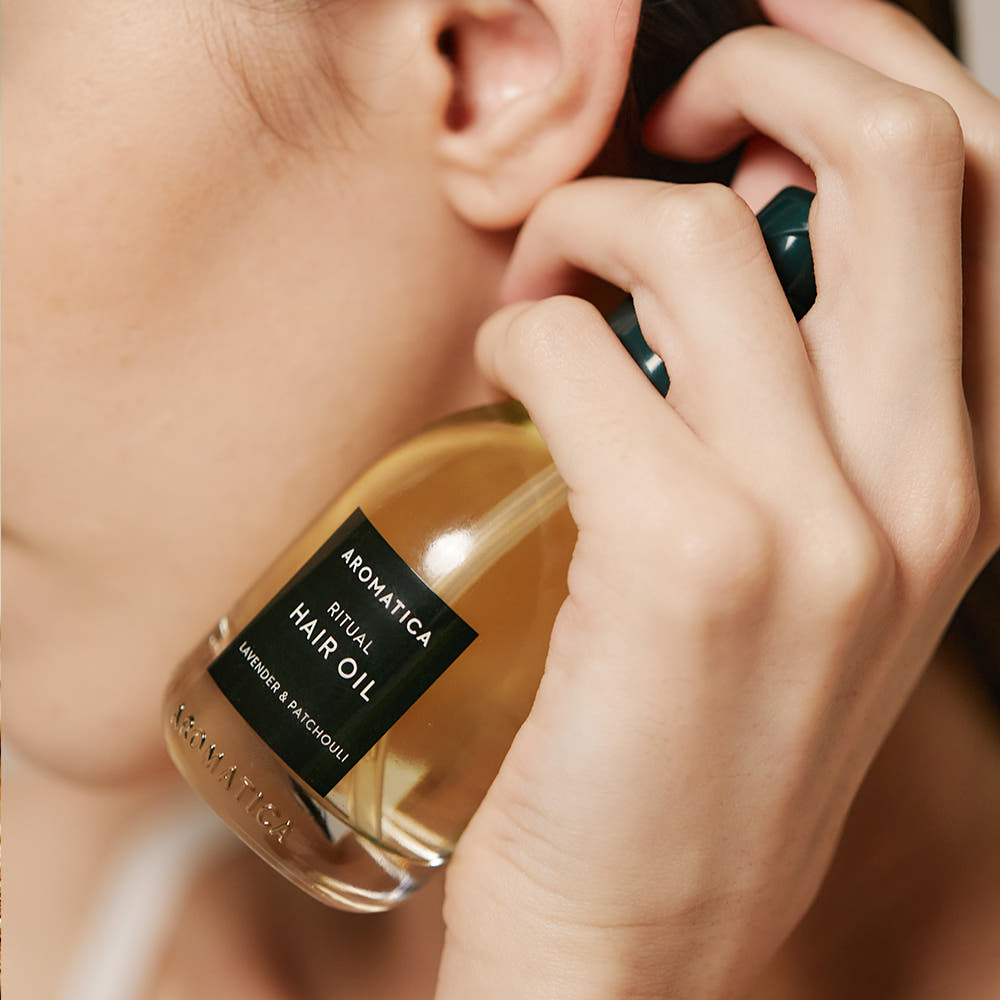 AROMATICA Ritual Lavender & Patchouli Hair Oil 50ml : la main applique l'huile sur cheveux, bouteille AROMATICA, visuel beauté.