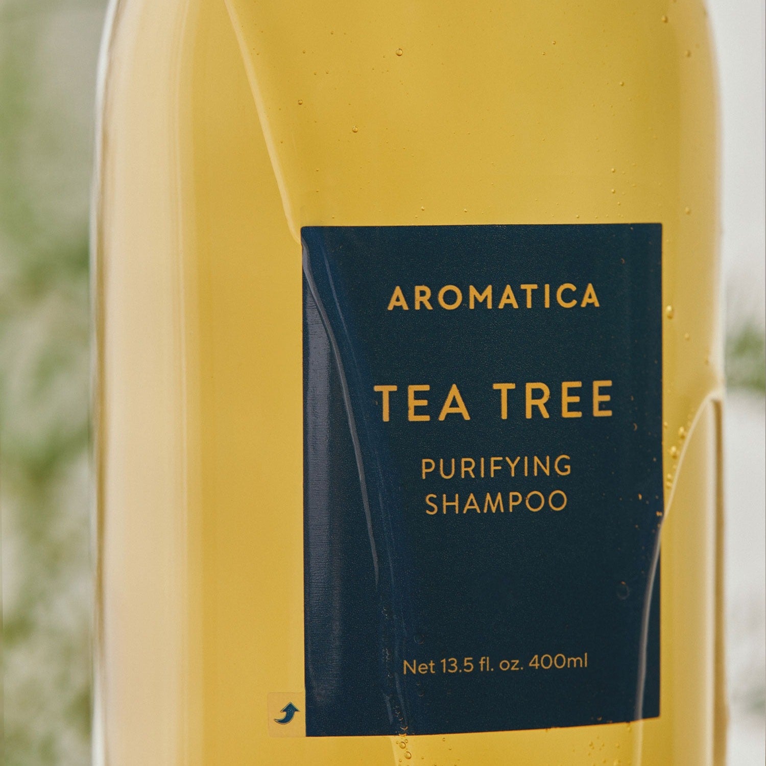 Décris l'image en français mentionnant AROMATICA Tea Tree Purifying Shampoo 750ML : flacon jaune avec étiquette Tea Tree, fond flou.