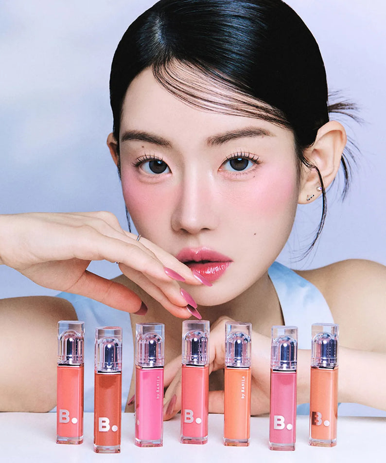 Femme utilisant les BANILA CO Dew Glow Tint pour un look maquillage frais et lumineux avec divers produits cosmétiques devant elle.