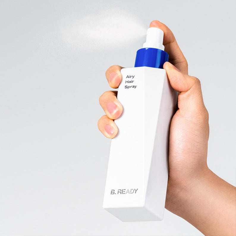Main tenant un flacon de BE.READY Airy Hair Spray 200ml, vaporisateur à la main, sur fond clair.