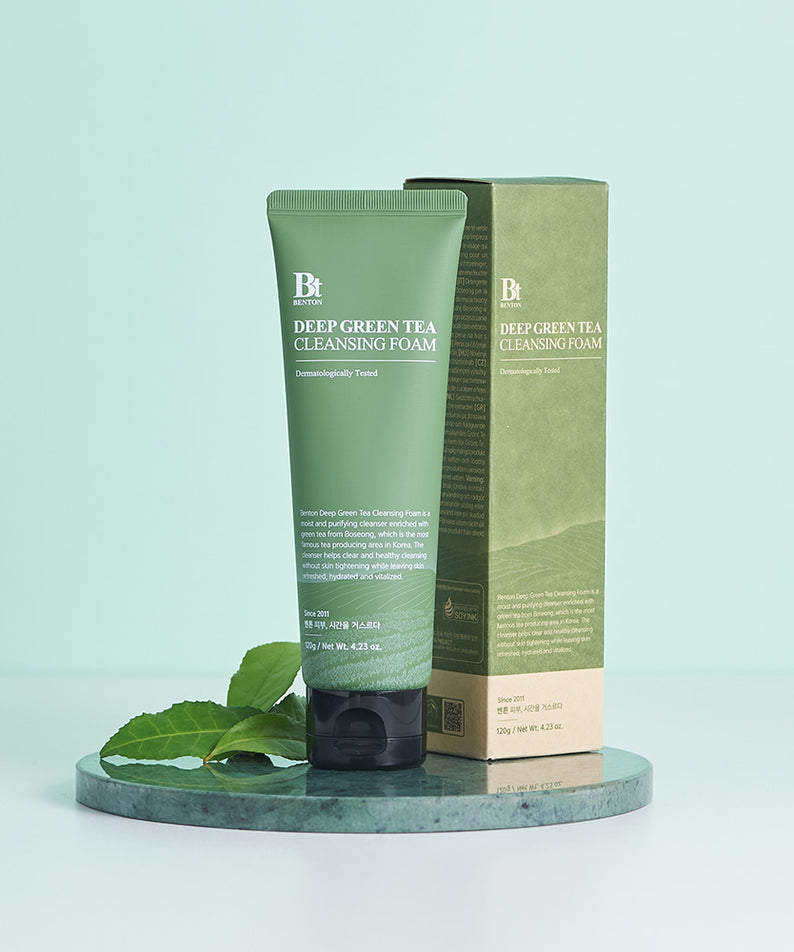 Décris l'image en français : BENTON Deep Green Tea Cleansing Foam 120g, flacon vert de la marque BENTON sur socle, feuilles de thé.
