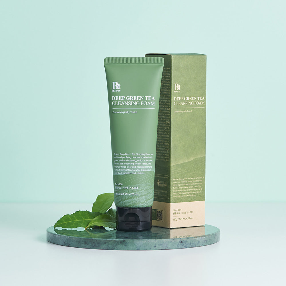 Décris l'image en français : BENTON Deep Green Tea Cleansing Foam 120g, flacon vert de la marque BENTON sur socle, feuilles de thé.