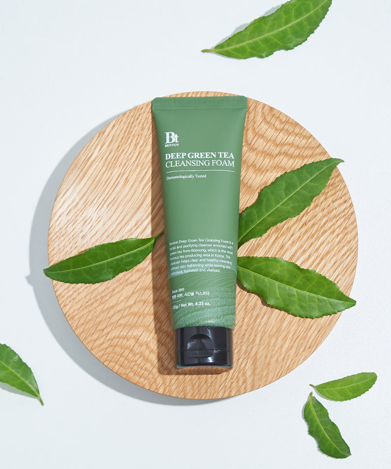Décris l'image en français : BENTON Deep Green Tea Cleansing Foam 120g, tube vert de la marque BENTON sur fond bois, feuilles de thé.