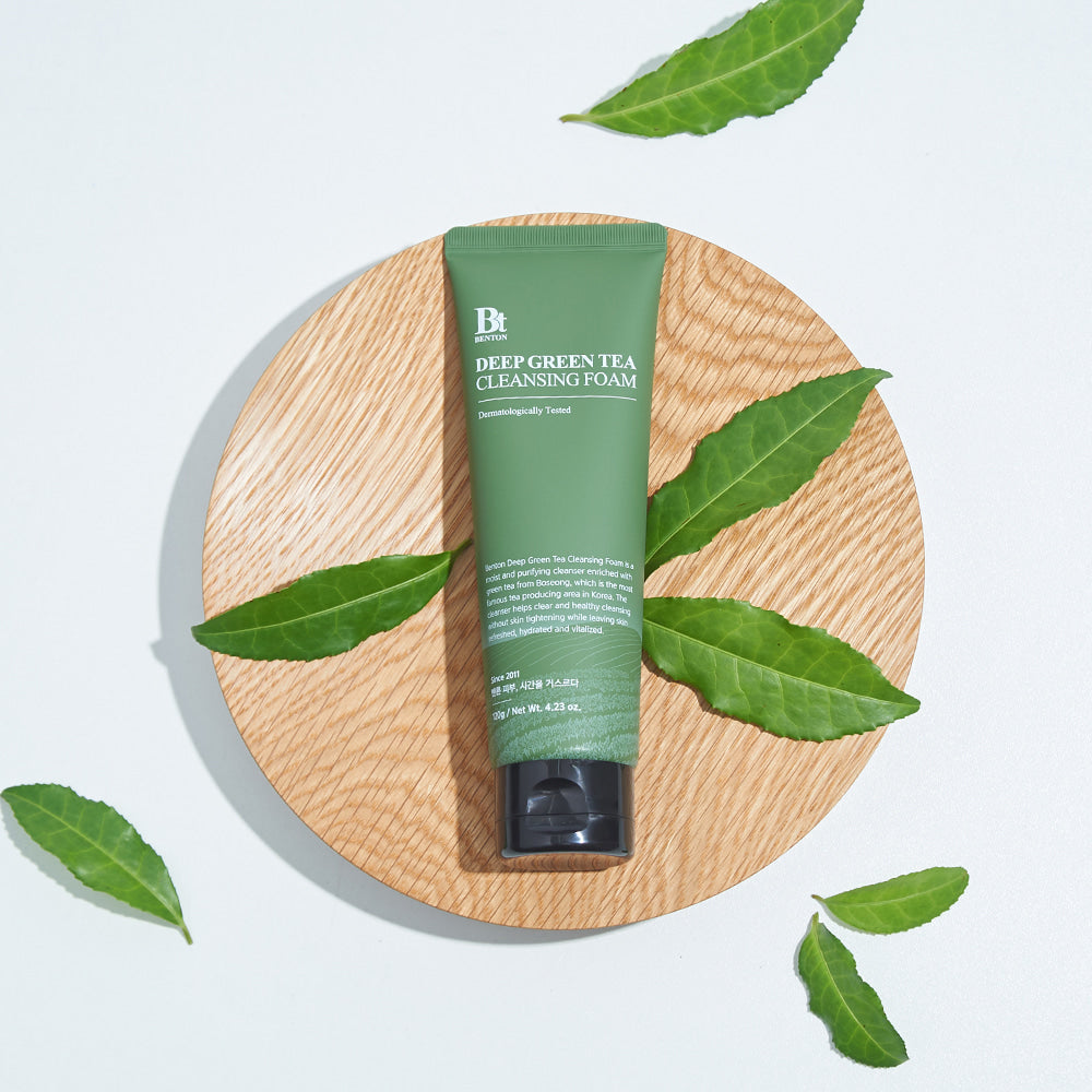 Décris l'image en français : BENTON Deep Green Tea Cleansing Foam 120g, tube vert de la marque BENTON sur fond bois, feuilles de thé.