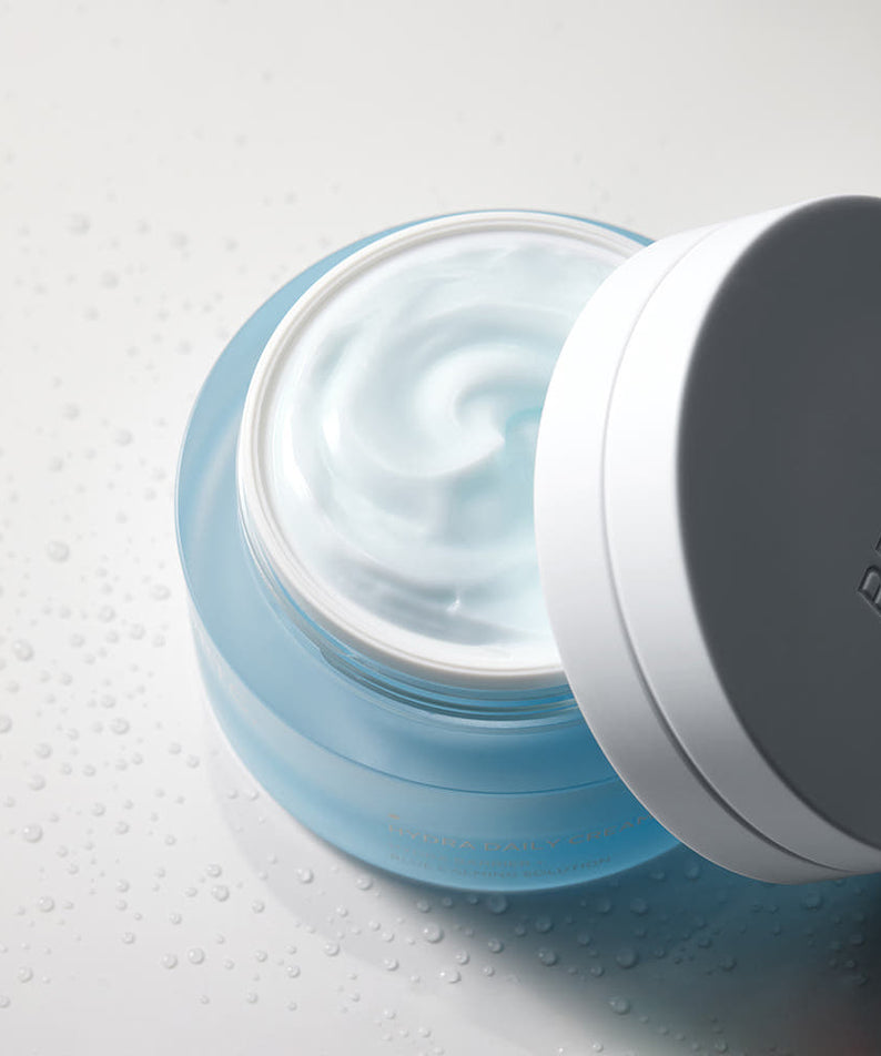 BRTC Hydra Daily Cream 50ml de BRTC, crème hydratante bleue en pot ouvert sur fond blanc avec gouttes d’eau.