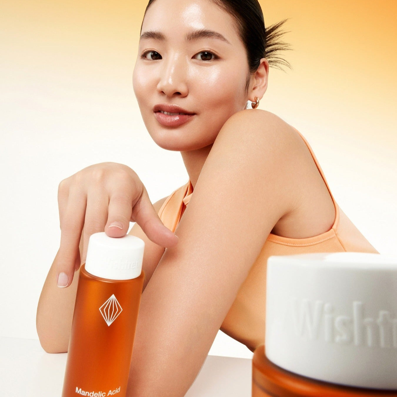 BY WISHTREND Madelic Acid Gentle Exfloliating Toner 150ml, femme en maquillage, flacon orange sur fond chaleureux