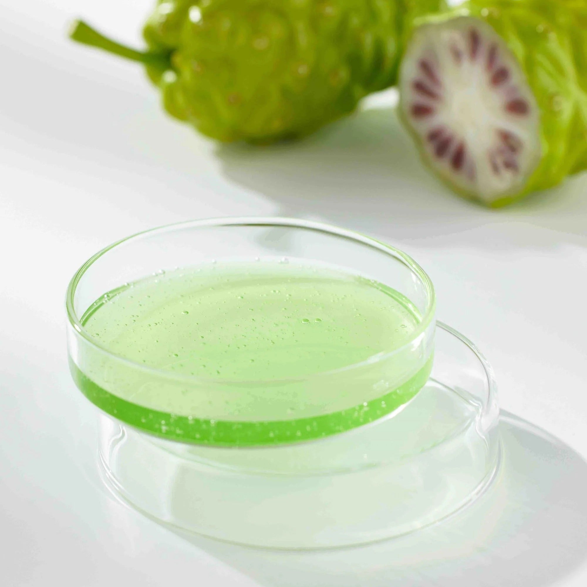 CELIMAX Noni Ampoule Calming+Radiance 100ml, flacon sur table blanche, gel vert et fruits de noni en arrière-plan.