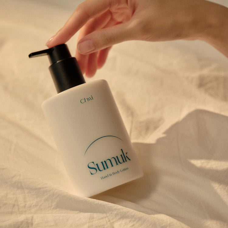 Main appliquant CHWI (CHI) Sumuk Hand & Body Lotion 300ml, flacon sur tissu clair, marque CHI visible, ambiance douce.