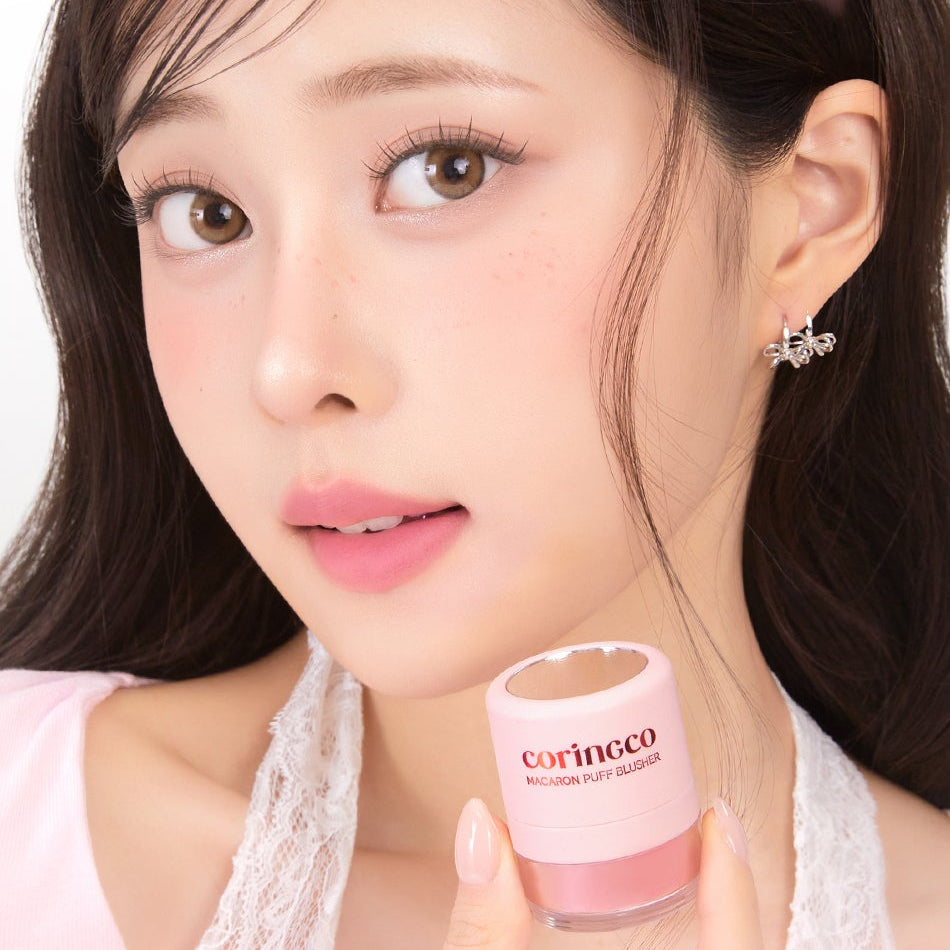 Décris l'image en français : CORINGCO Macaron Puff Blusher, marque Coringco, sur visage maquillé, main tenant le produit.