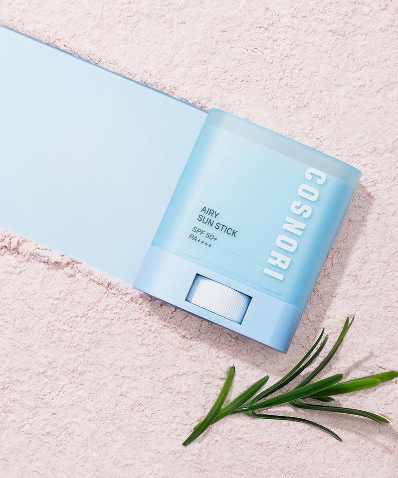 COSNORI Airy Sun Stick 20.5g (SPF50+ PA++++) sur fond pastel, vue produit et herbes, ambiance été ensoleillée