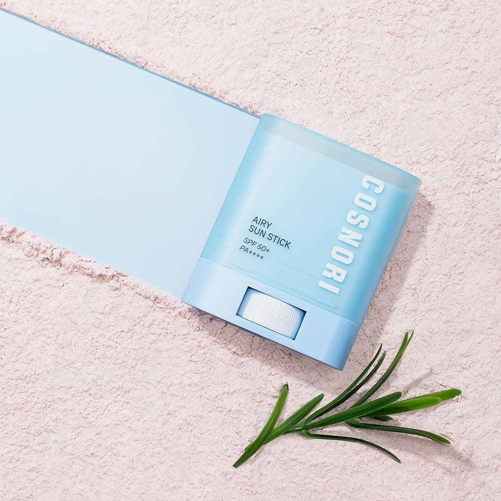 COSNORI Airy Sun Stick 20.5g (SPF50+ PA++++) sur fond pastel, vue produit et herbes, ambiance été ensoleillée