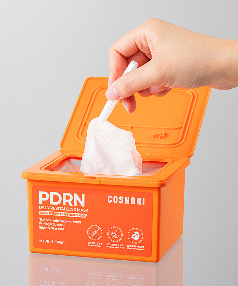 Main tenant COSNORI PDRN Daily Revitalizing Mask 30 sheets, boîte orange avec les lingettes masques PDRN, contexte cosmétique