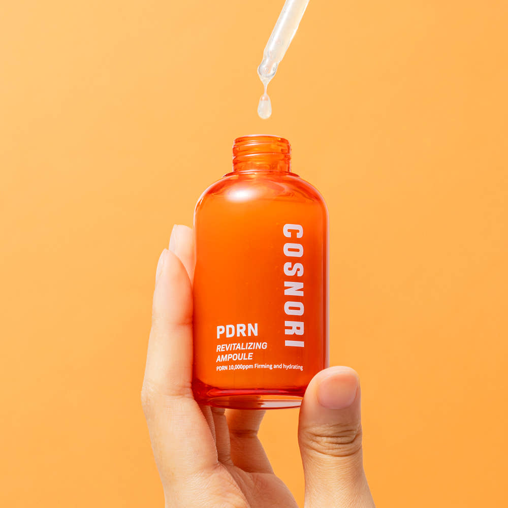 COSNORI PDRN Revitalizing Ampoule 50ml : main tenant le flacon orange avec pipette, fond orange, contexte soin beauté