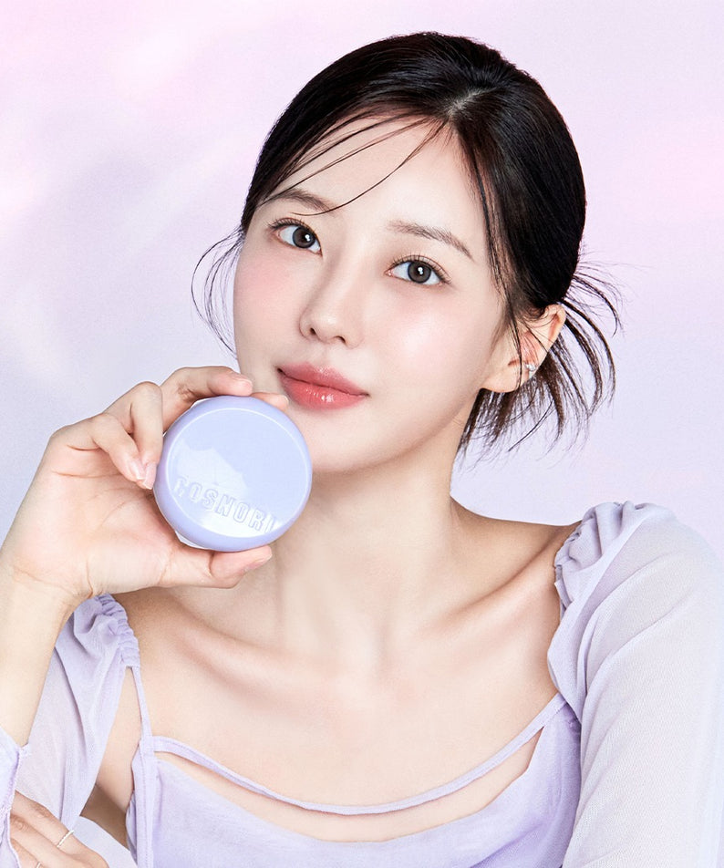 Jeune femme utilisant COSNORI Panthenol Barrier Cushion SPF50+PA++++, produit de soin à écran solaire, fond mauve, peau lumineuse.