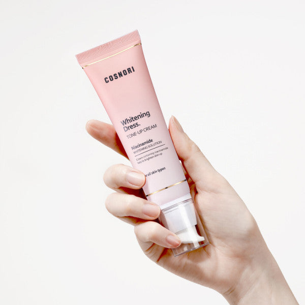Main tenant COSNORI Whitening Dress Tone-Up Cream 50ml, tube rose sur fond clair, produit de beauté blanc/doré.