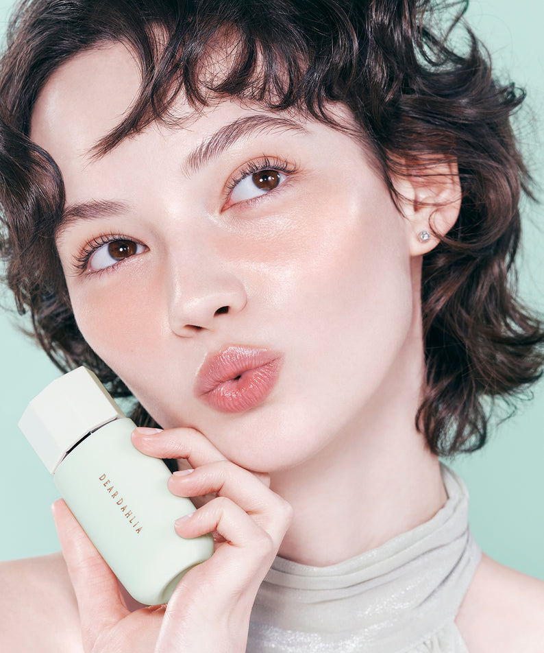 Femme posant avec le produit DEAR DAHLIA Prime Layer Correcting Primer, illuminant un teint éclatant et naturel.