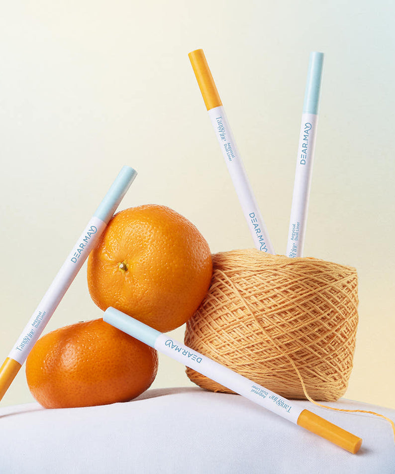 DEAR MAY Tangerine Aegyo-sal Dual Liner de la marque DEAR MAY, stylos maquillage près d’oranges et de filet à la lumière.