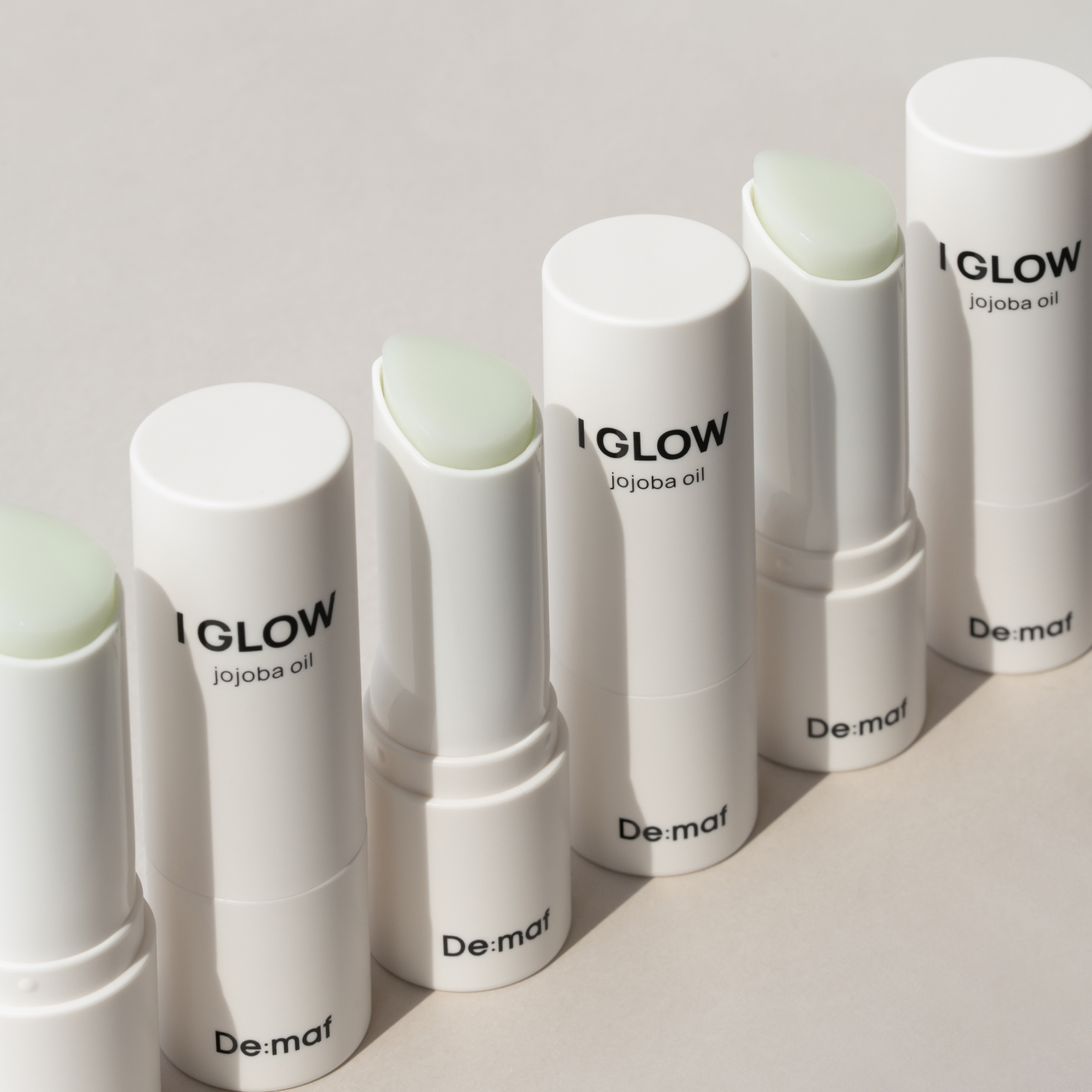 Décris l'image en français en mentionnant le produit, la marque et le contexte visuel: DE:MAF Eye Glow Jojoba Oil Multi Balm 9g, baumes tubes blancs sur fond clair.