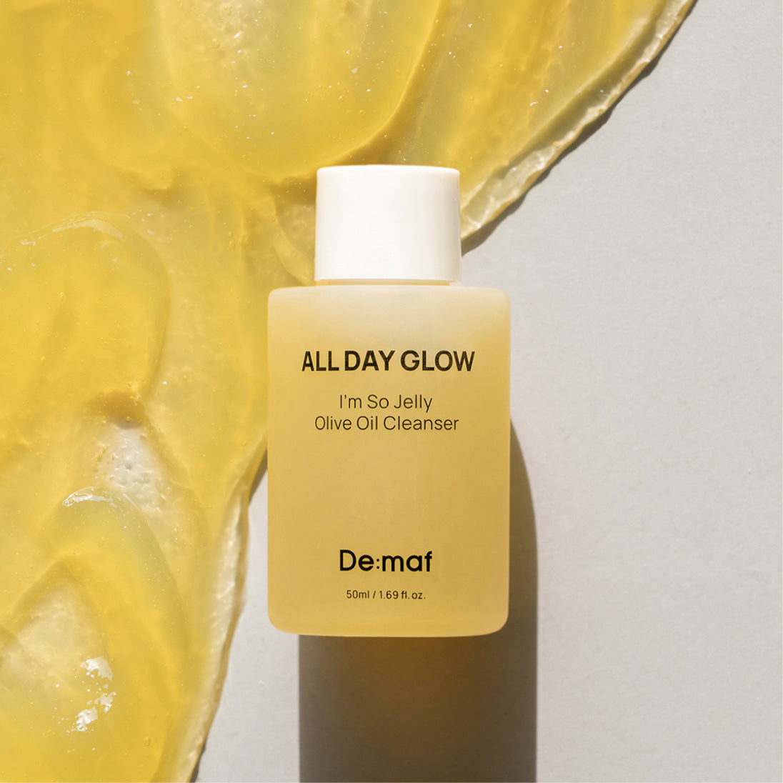 DE:MAF I'm So Jelly Olive Oil Cleanser 50ml De:màf, flacon sur fond jaune façon gelée, texture d’olive en arrière-plan