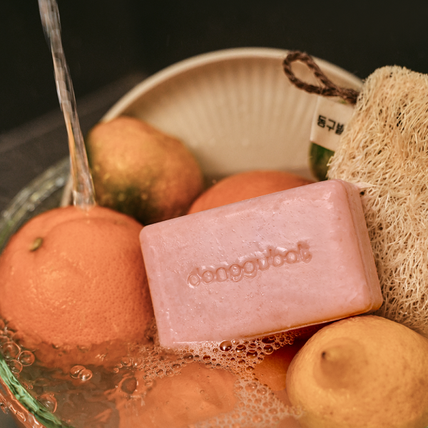DONGGUBAT Blooming Jaungo Washing Bar 80g, savon rose de la marque, posé dans une bassine avec fruits et brosse, ambiance douche.