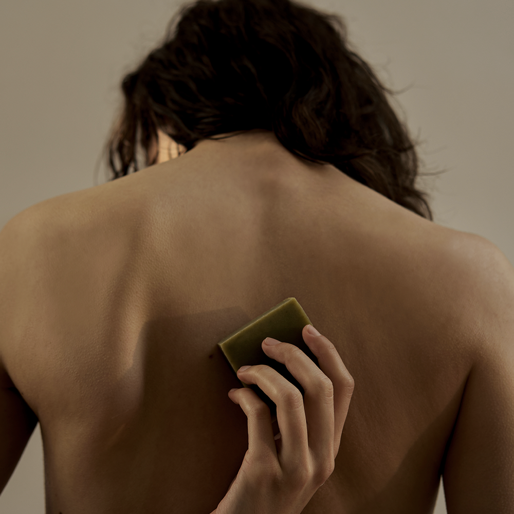 Décris l'image en français : DONGGUBAT Dream Soap Lattuce, marque DONGGUBAT, tenue nue dos, savon vert sur la peau.