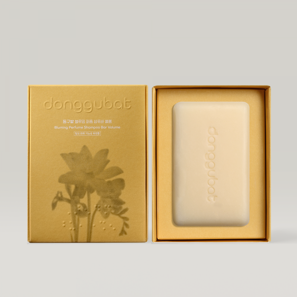 Décris l'image en français : DONGGUBAT Field Blooming Perfume Shampoo Bar Volume 80g, savon dans sa boîte jaune, sur fond clair.