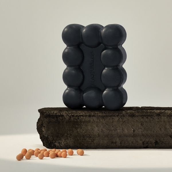 DONGGUBAT Gwalsa Therapy Scrub Shampoo Bar Geocharcoal 95g, barre noire sur bûche bois et billes, fond clair.