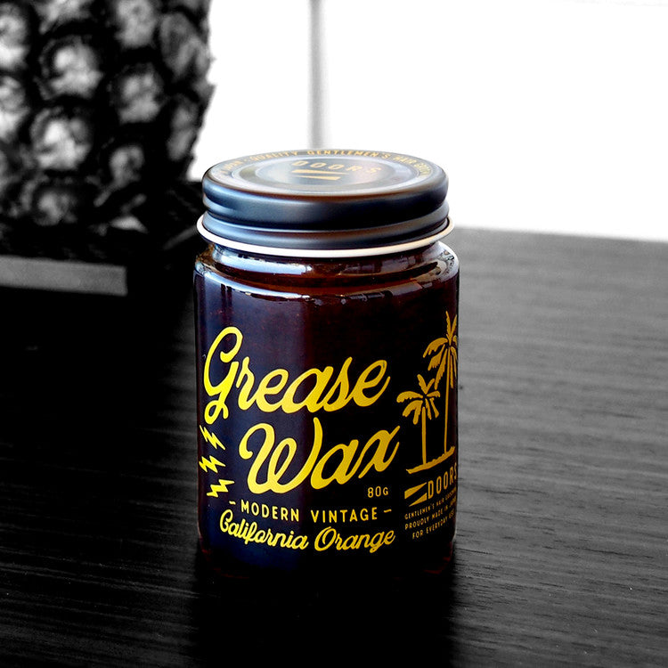 DOORS California Beach Orange Grease Wax 80g, pot sur table, ambiance plage californienne, couleur orange et marque visible.