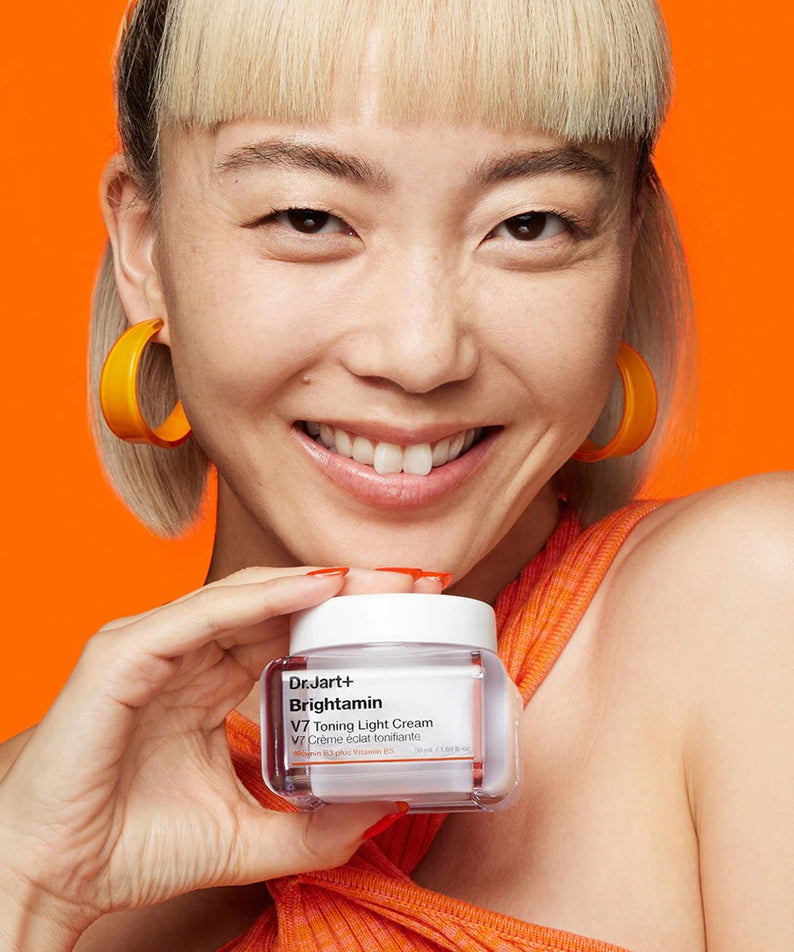 Femme souriante tenant un pot de crème DR.JART+ Brightamine V7 Toning Light Cream sur fond orange vif.