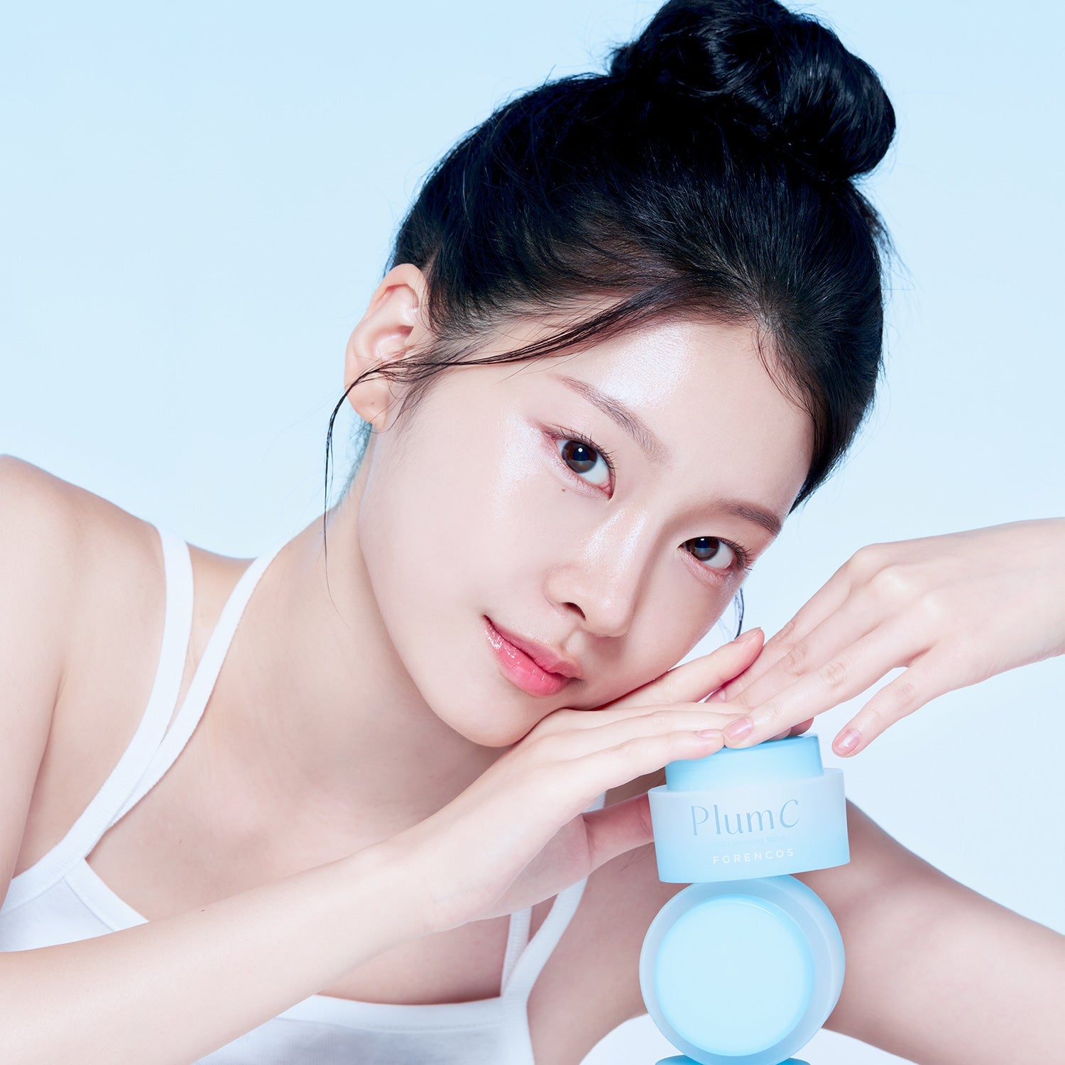 Décris l'image en français : FORENCOS Plum C Mild Cleansing Balm 50ml, pot et baume sur peau, modèle beauté en studio bleu.