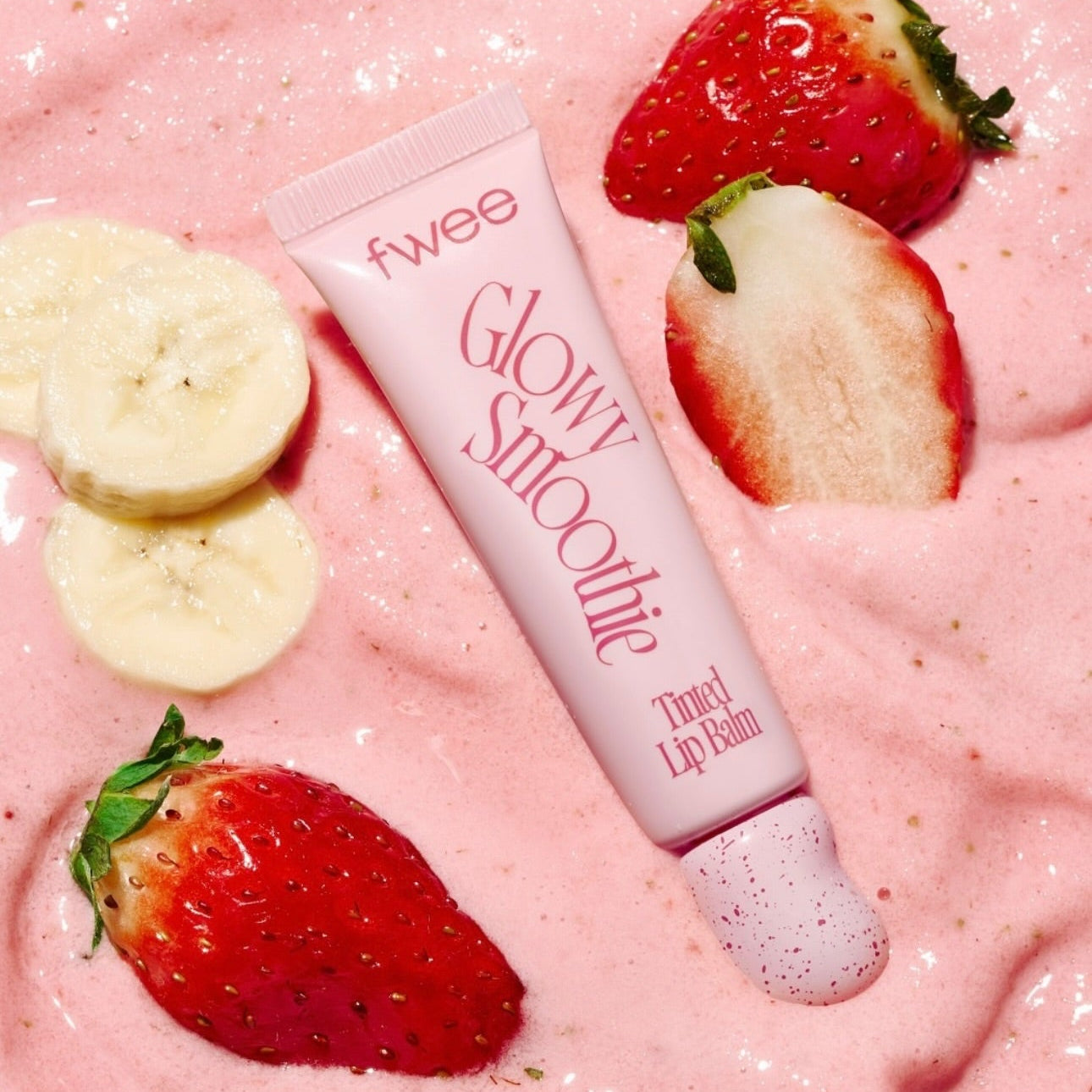FWEE Glow Smoothie Tinted Lip Balm de la marque FWEE, sur fond rose avec fraises et tranches de banane.