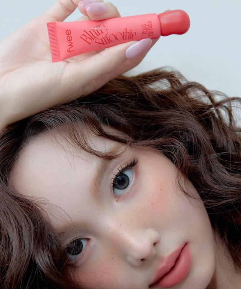 FWEE Glow Smoothie Tinted Lip Balm de la marque FWEE, baume teinté rose sur visage, main en gros plan, contexte beauté