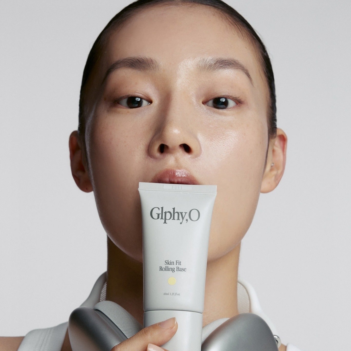 Décris l'image en français : GLPHYO Skin Fit Rolling Base 40ml de GlphyO, tube crème tenu sur fond clair, visage maquillé