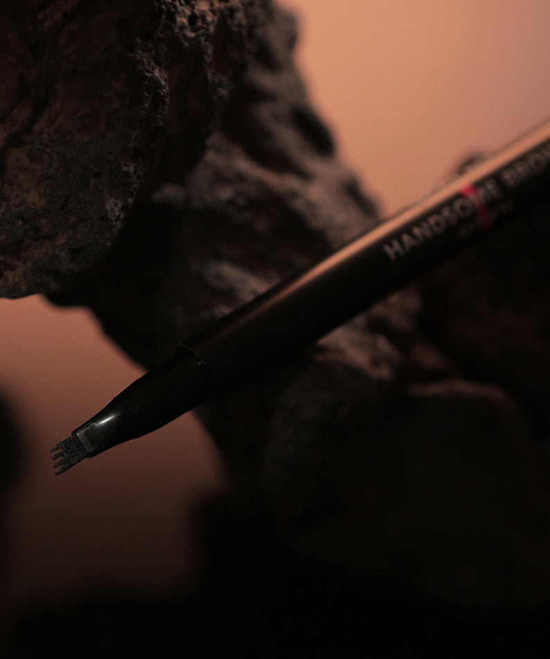 GRAFEN  Handsome Tattoo Brow 0.8g : crayon sourcils noir sur fond maquillage, main tenant le produit, vue rapprochée