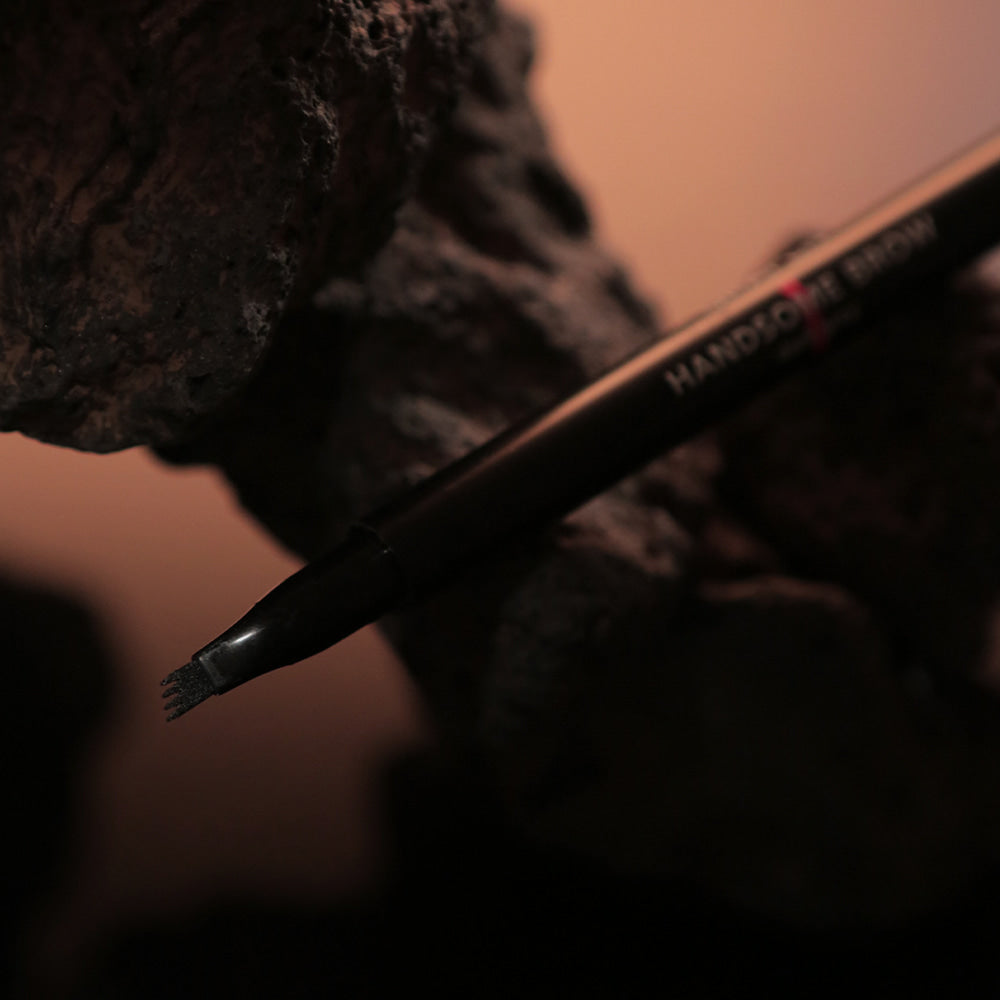 GRAFEN  Handsome Tattoo Brow 0.8g : crayon sourcils noir sur fond maquillage, main tenant le produit, vue rapprochée