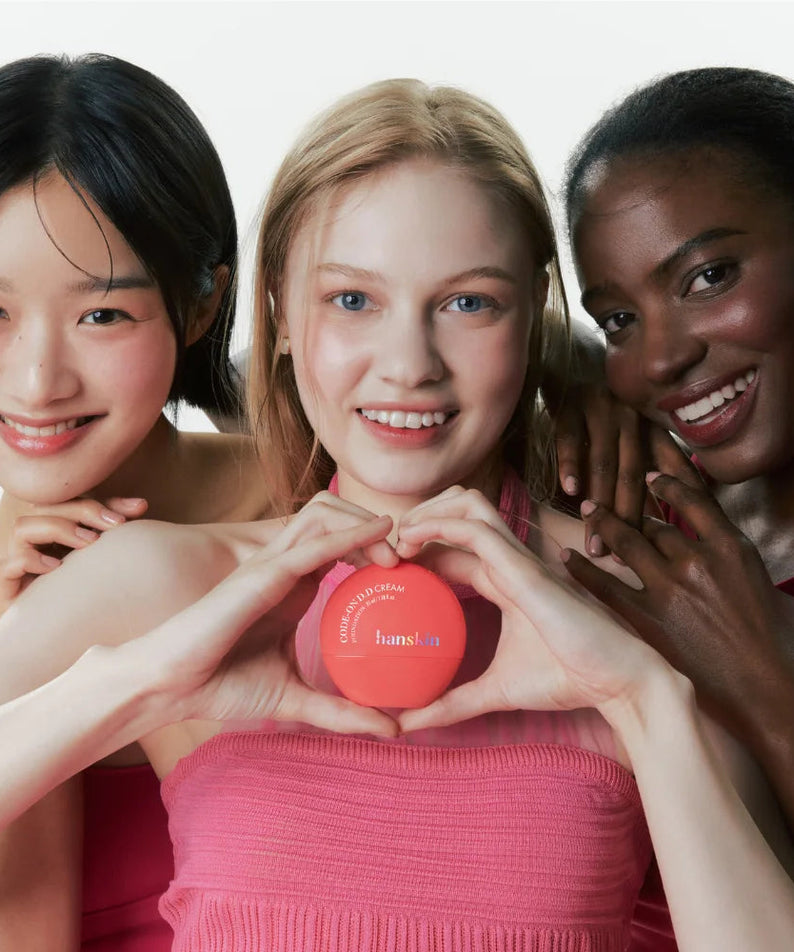 Trois femmes souriantes tenant HANSKIN Code On DD Cream Foundation 35ml (boîtier rouge) en fond studio, marque HANSKIN.