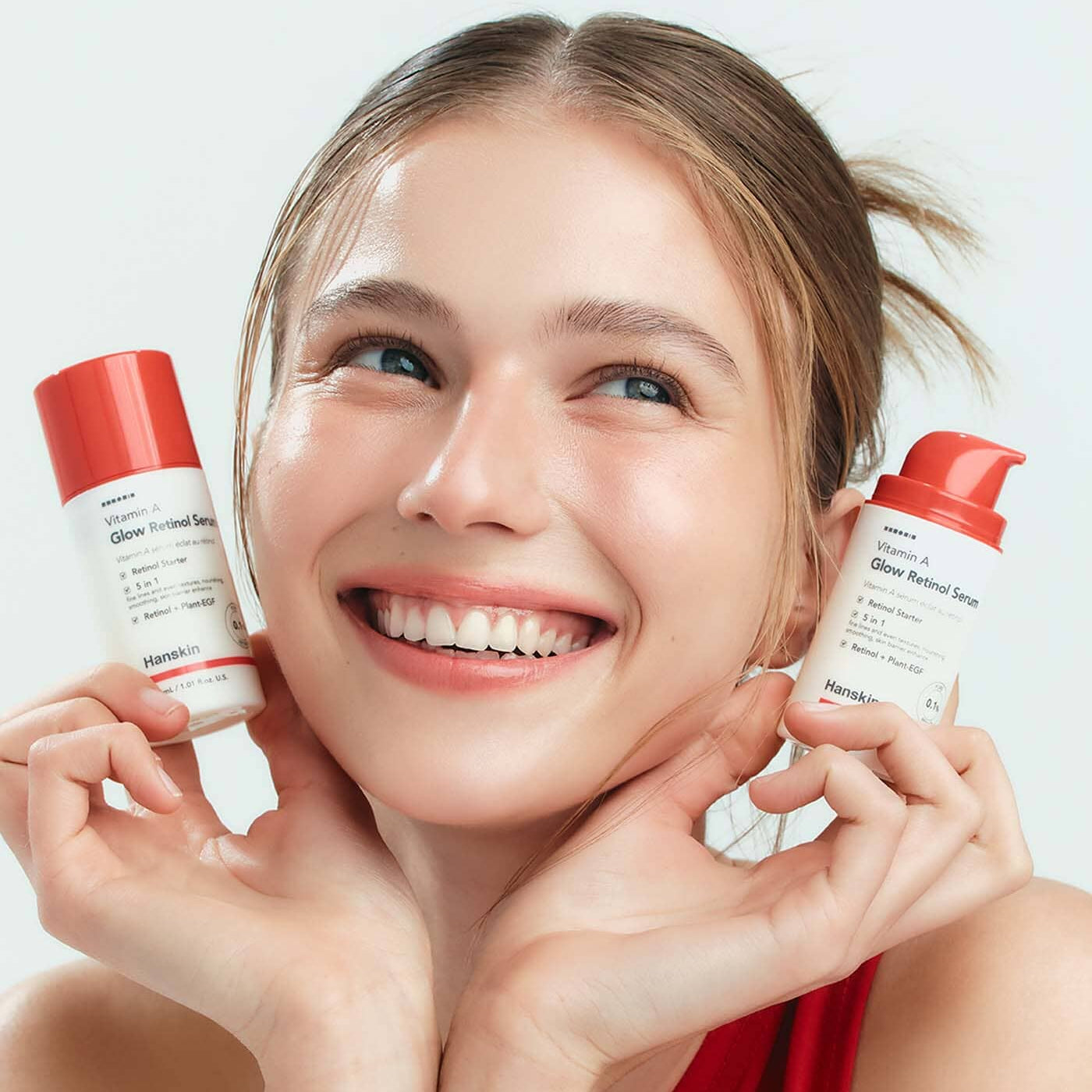 Décris l'image en français : HANSKIN Vitamin A Glow Retinol Serum 30ml, marque HANSKIN, femme souriante tient deux flacons.