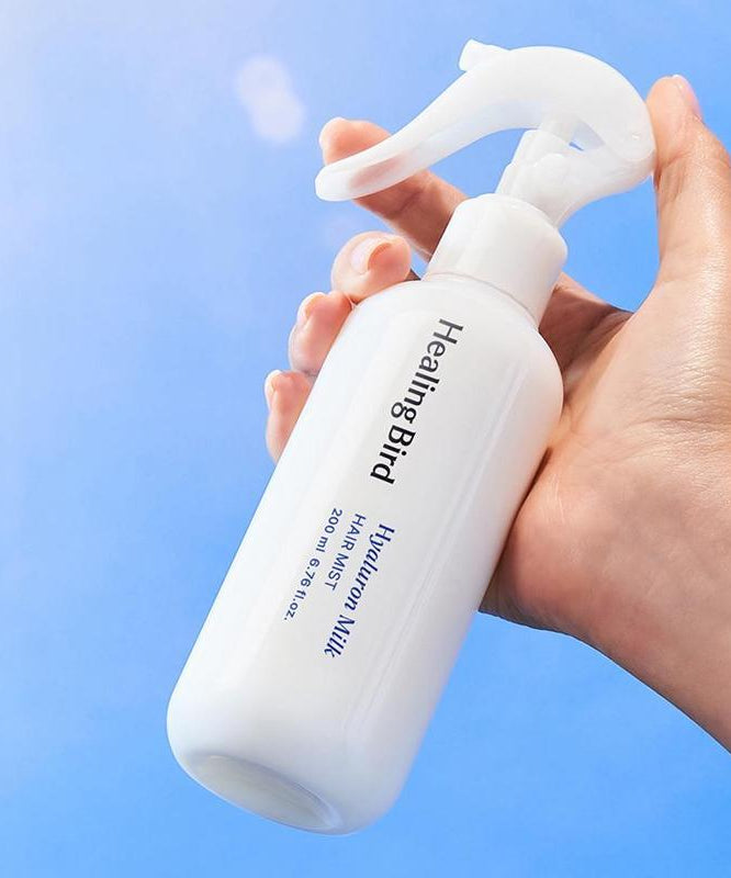Bouteille de HEALING BIRD Hyalutonic Milk Hair Mist 200ml tenue dans une main, sur fond bleu.