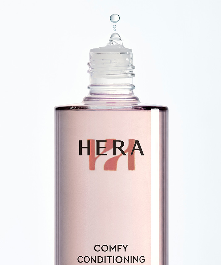 HERA  Comfy Conditioning Essence 150g, flacon cosmétique rose sur fond blanc, marque HERA en gros plan.