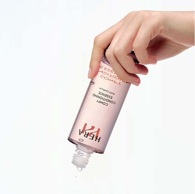 Main tenant HERA  Comfy Conditioning Essence 150g, flacon cosmétique sur fond blanc, contexte soin beauté en français.