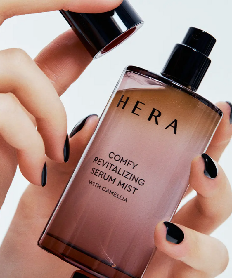 Décris l’image en français : HERA Comfy Revitalizing Serum Mist 90ml de la marque HERA, mains appliquant la brume sur flacon.