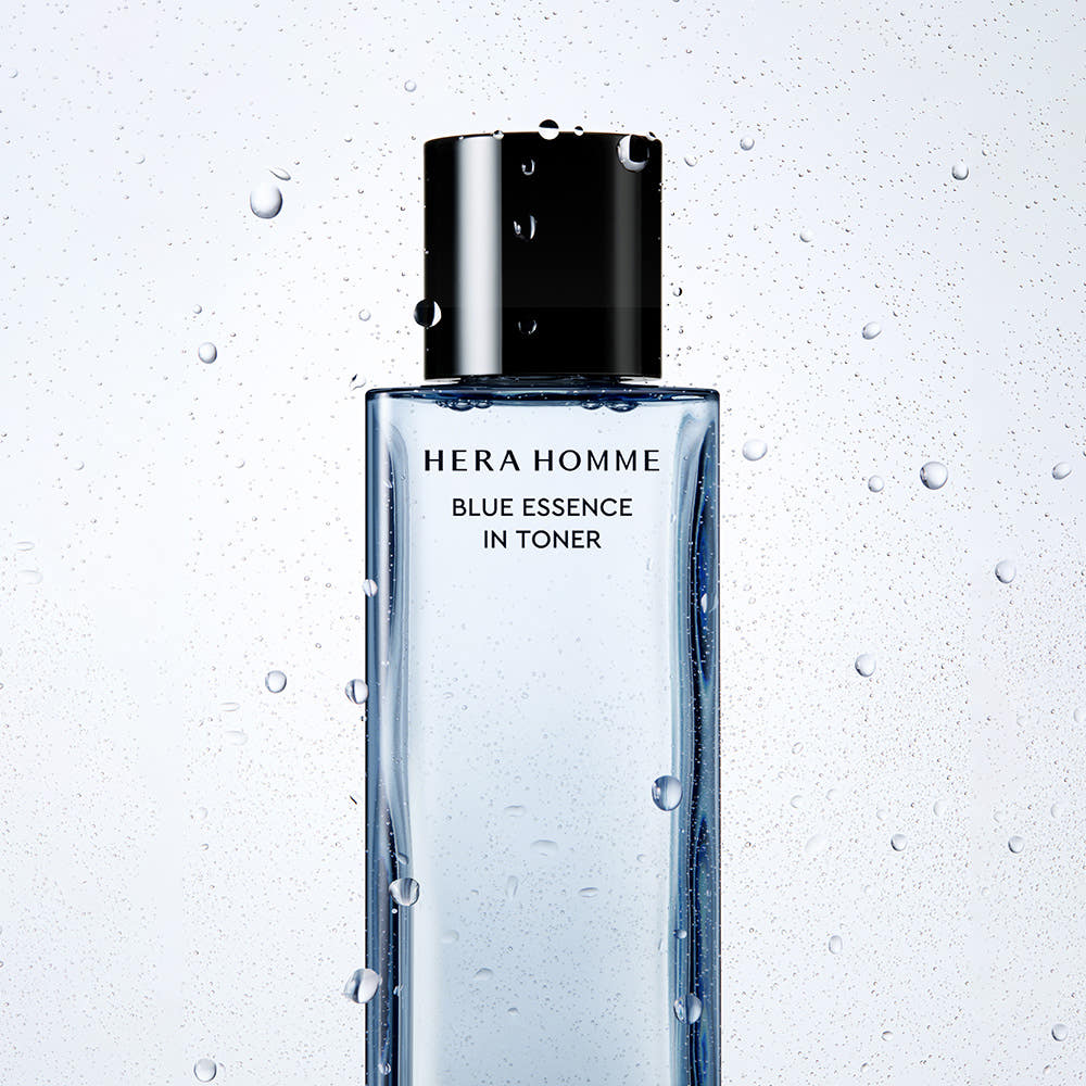 HERA  Homme Blue Essence In Toner 125ml : flacon noir/bleu sur fond clair avec gouttes d’eau, design salle de bain.