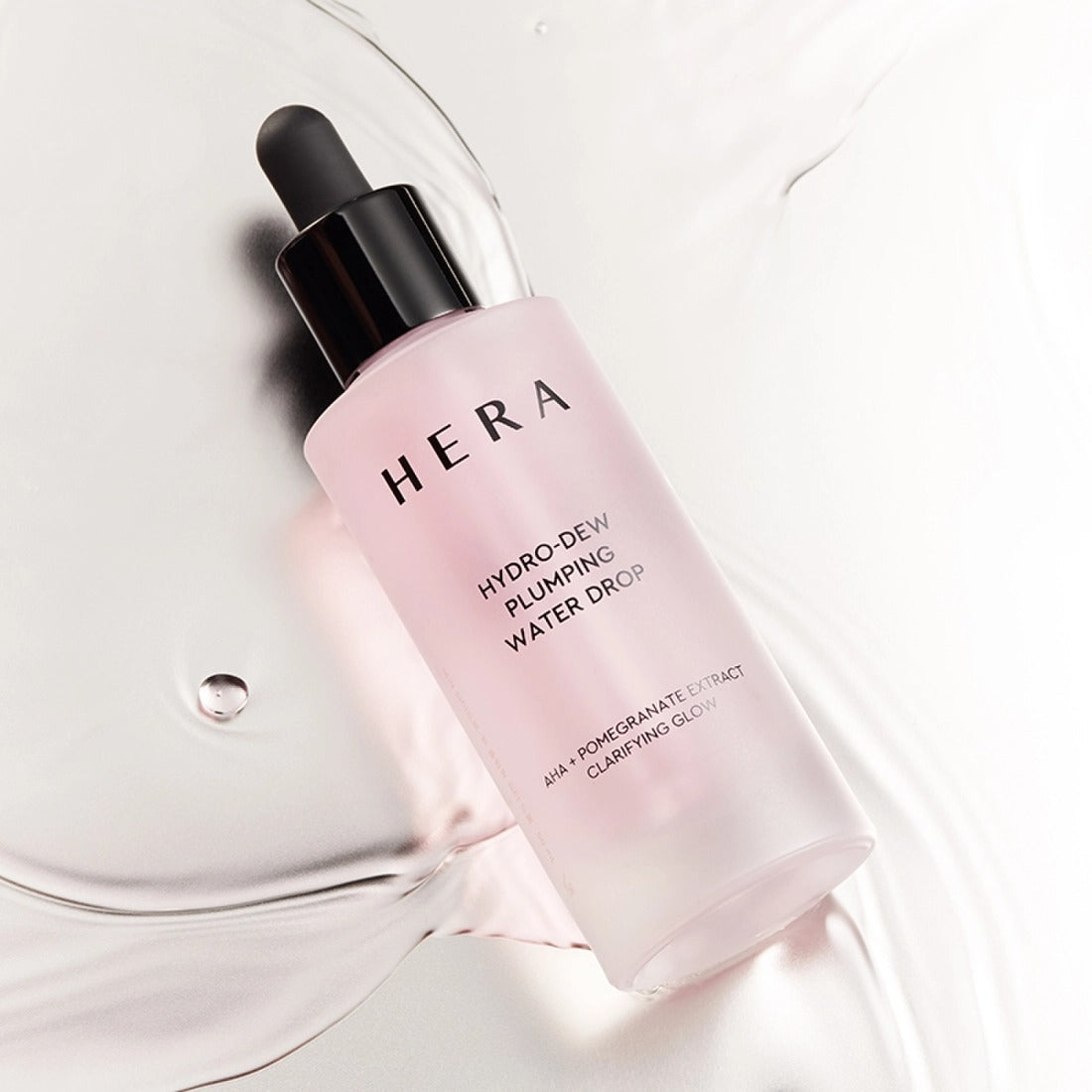 Décris l'image en français : HERA Hydro-Dew Pluming Water Drop 50ml, flacon HERA sur fond blanc, éclaboussures d’eau.