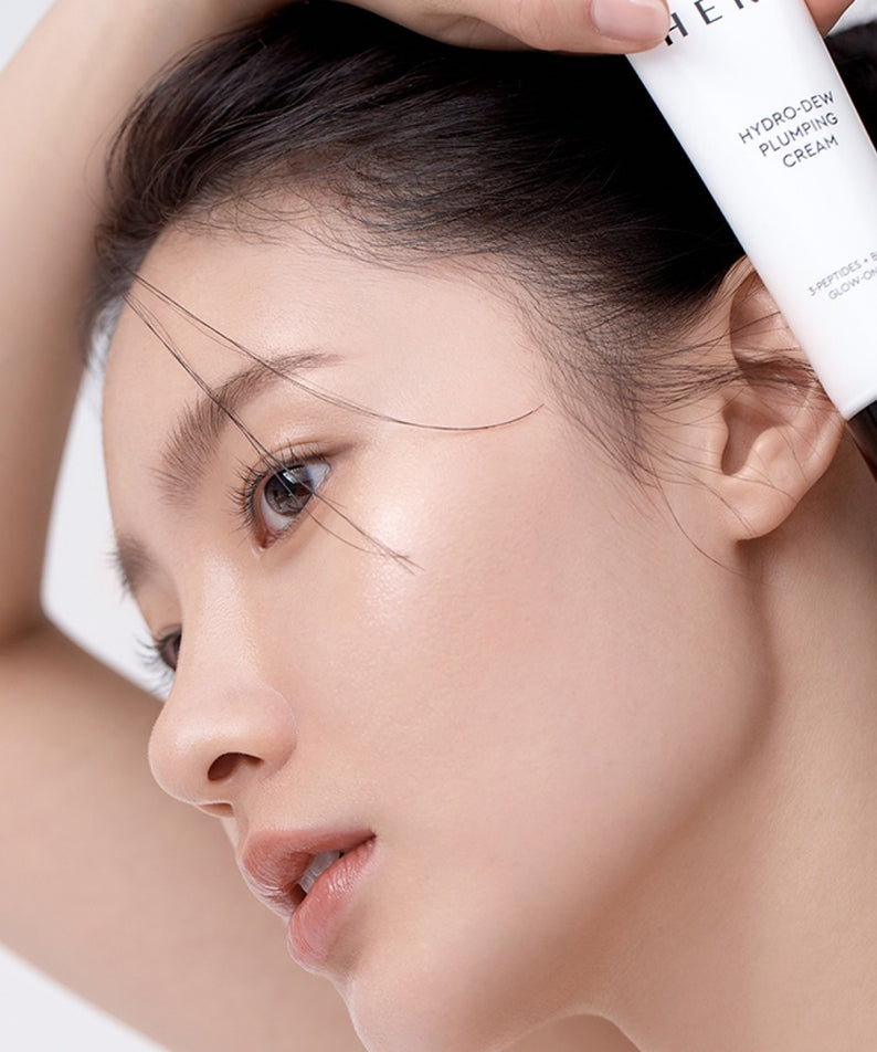 HERA Hydro-Dew Plumping Cream 50ml, gros plan sur un visage, tube de crème HERA sur la main, contexte skincare en studio