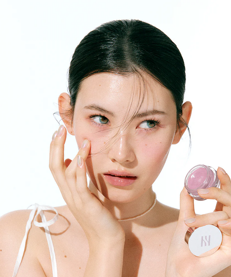 HERA Sensual Lip & Cheek 7g : maquillage corail sur visage lumineux, modèle appliquant avec le produit HERA, fond clair.