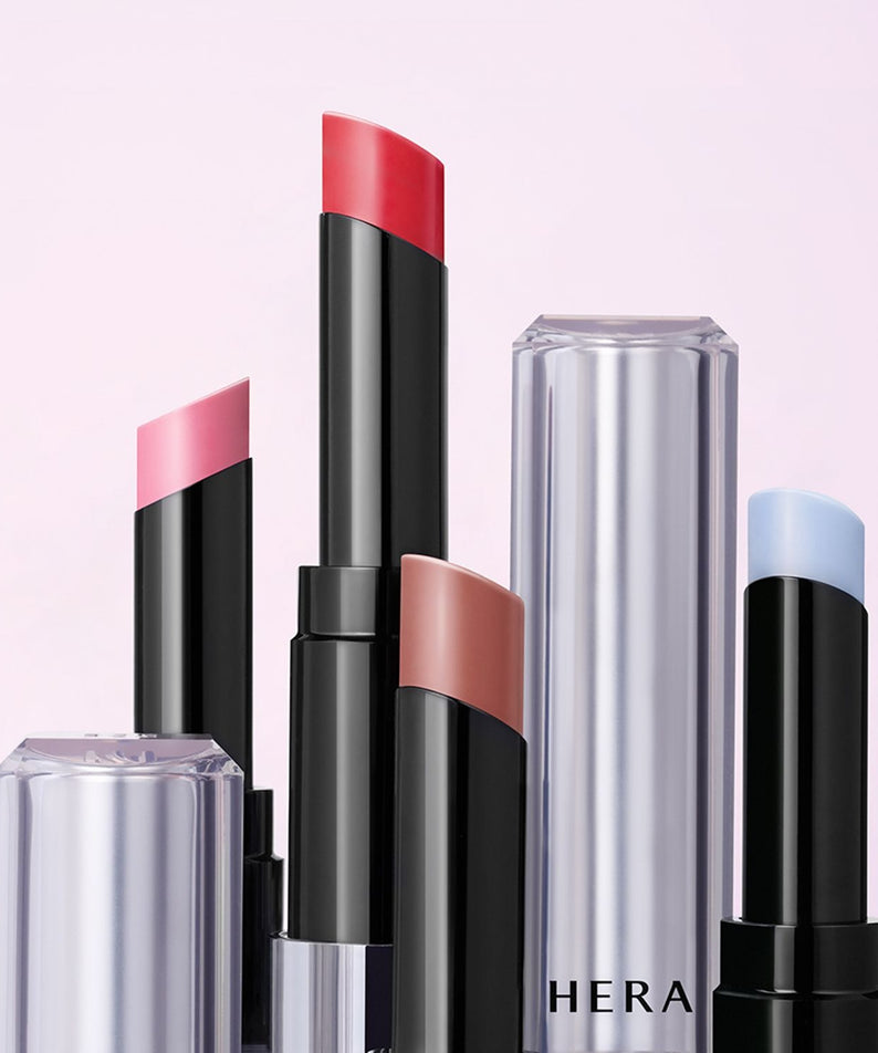 Décris l'image en français: HERA Sensual Nude Balm 3.5g, rouge à lèvres nude HERA sur fond rose, pack noir et tube transparent.