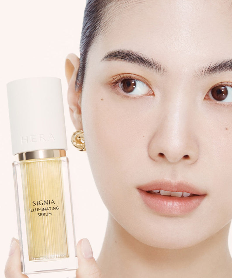 Décris l'image en français : HERA  Signia Illuminating Serum 30ml de HERA, flacon doré tenu par un visage maquillé sur fond clair.