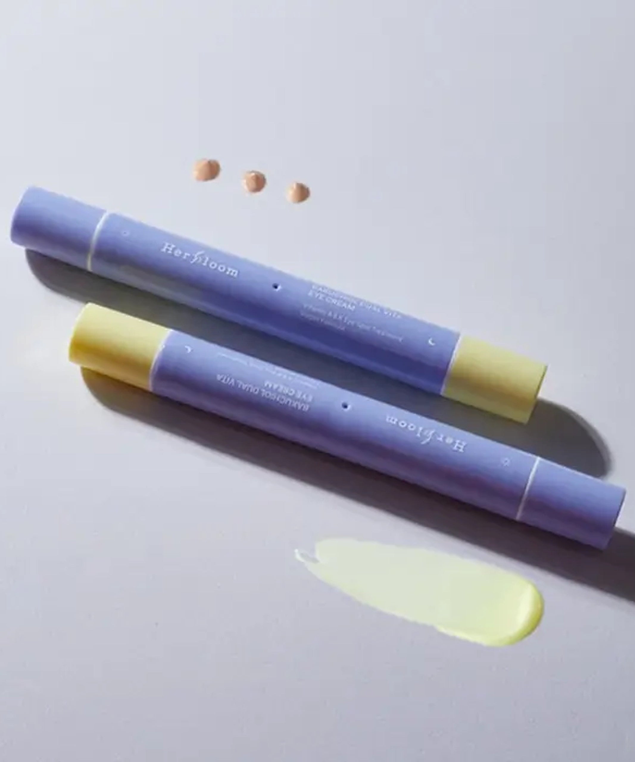 Décrit l’image en français : HERBLOOM  Bakuchiol Dual Vita Eye Cream, tube bleu-violet, texture crème près d’un fond clair.