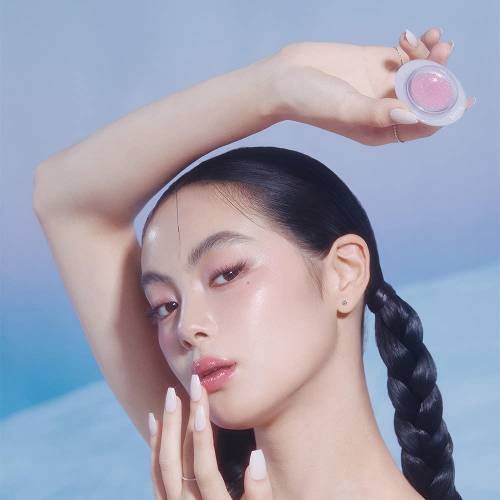 HINCE Low Glow Dewy Ball (Lip & Cheek) de HINCE, main tenant le baume dégradé sur fond bleu ciel, visage maquillé.